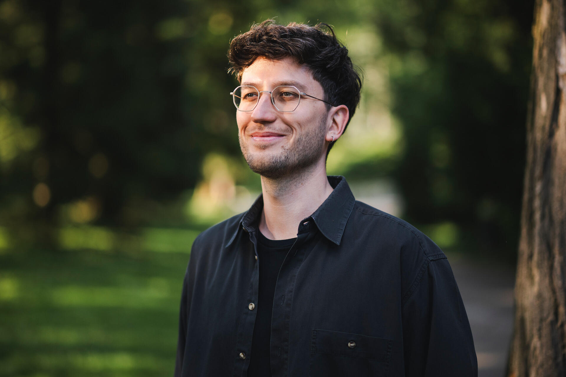 Jakob Bouchal, der ein schwarzes Hemd und eine Brille trägt, steht vor einem Hintergrund satter, grüner Blätter und Pflanzen, und lächelt.