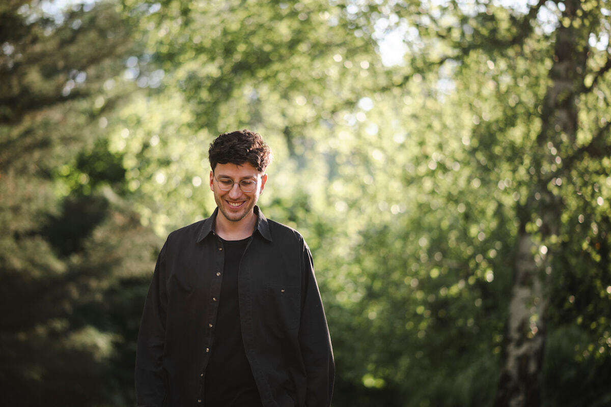 Jakob Bouchal, der ein schwarzes Hemd trägt, steht vor einem hellen Hintergrund grüner Blätter und Bäume, und lächelt.