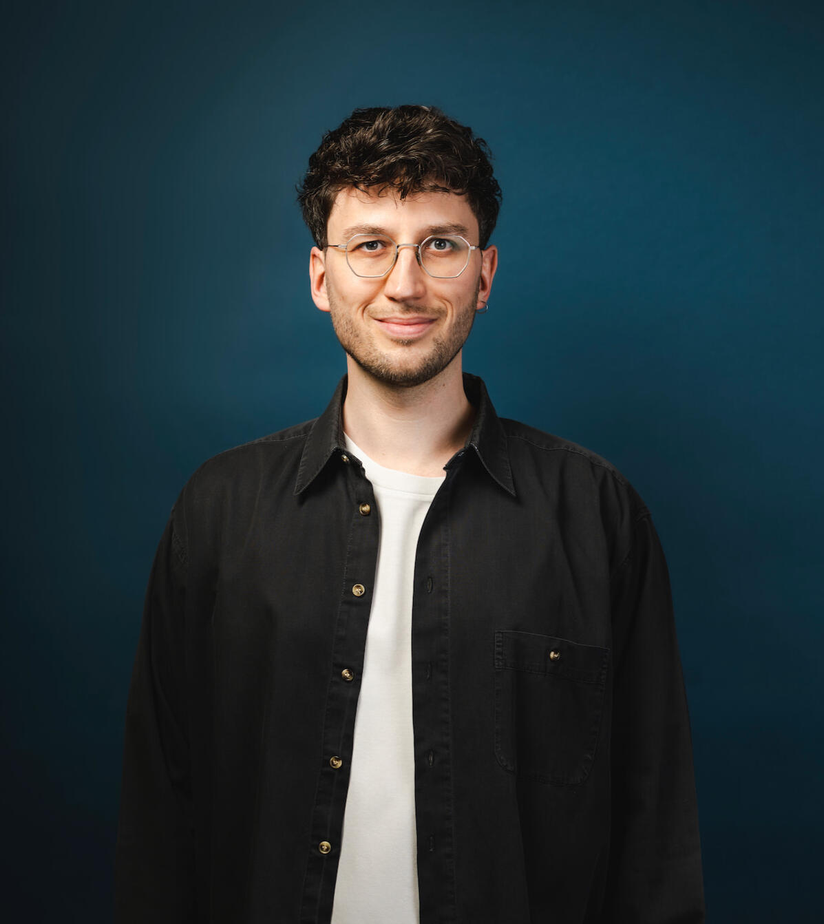 Jakob Bouchal, der ein schwarzes Hemd und ein weißes T-Shirt trägt, steht vor einem dunkleblauen Hintergrund und lächelt.