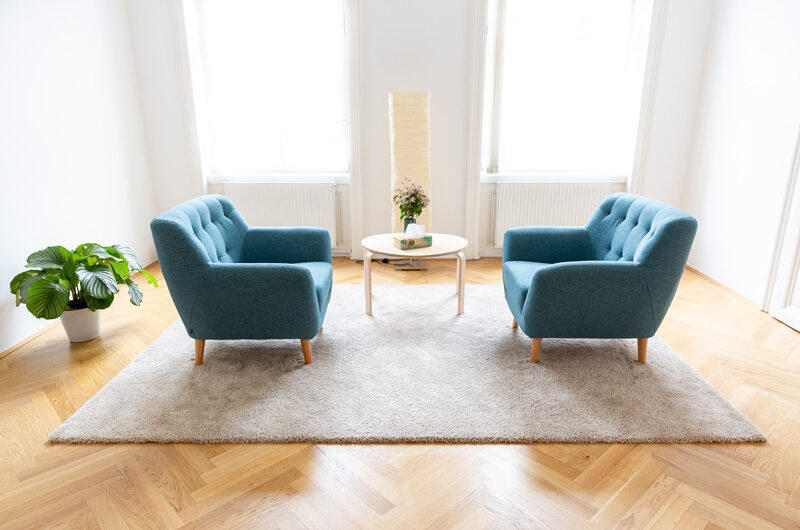 Zwei blaue Polstersessel stehen links und rechts von einem weißen runden Tisch auf einem hellen Teppich. Auf der linken Seite steht eine Pflanze, und in der Mitte des hellen Raums stehen eine hohe Lampe und eine Blumenvase vor großen Fenstern.